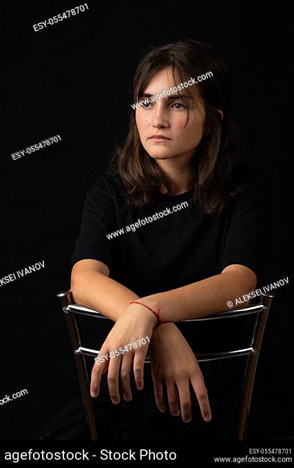 Portrait of a young girl sitting on a high chair and folded her hands on the back of a chair, black background