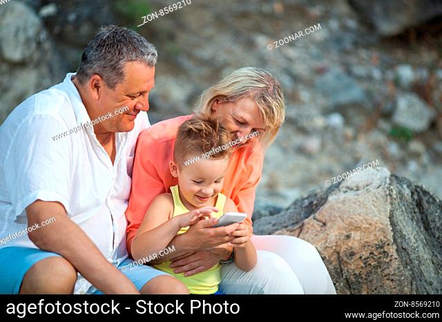 Happy grandparents and grandson with smartphone outdoor. Grandmother and grandfather watching the boy playing with mobile