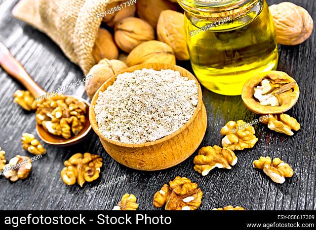 Walnut flour in bowl, nuts on a table, in a spoon and in a bag, oil in glass jar on black wooden board background