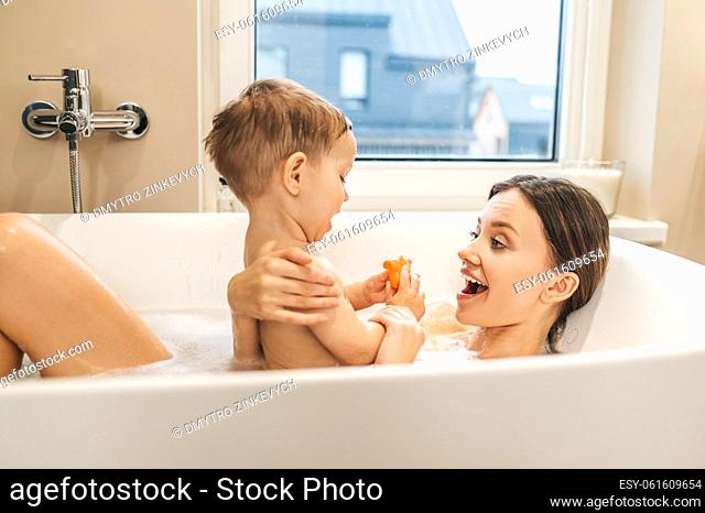 High-spirited young woman lying in the bath and supporting her baby boy seated on her