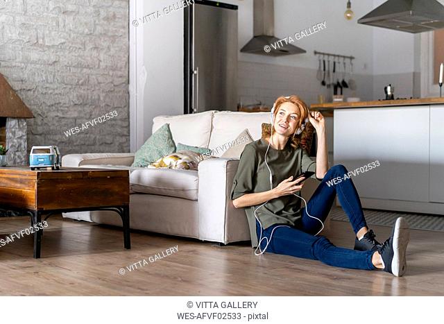 Young woman sitting on floor at home, using smartphone, wearing headphones