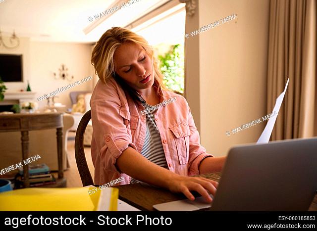 Caucasian woman sitting at table working in living room, using laptop and talking on smartphone