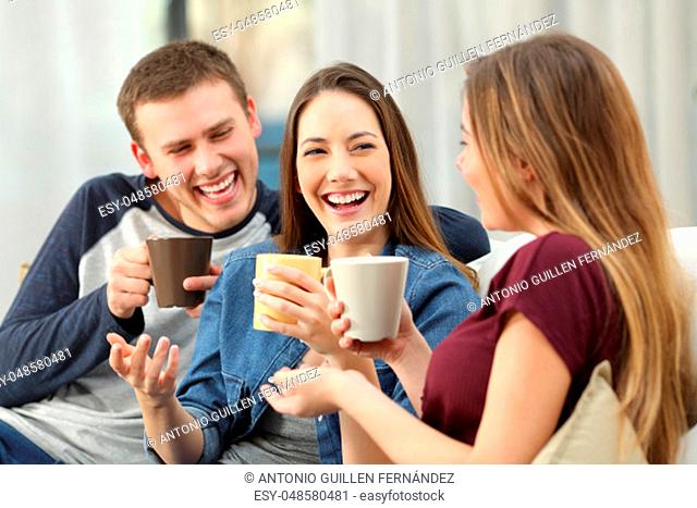 Three happy friends talking and laughing loud holding drinks sitting on a sofa in the living room at home