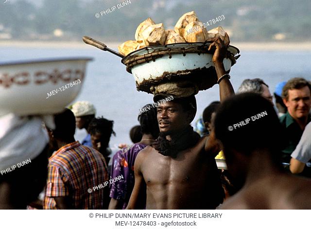 Coconut seller carries his wares on his head, on Goderich Beach, Sierra Leone, West Africa