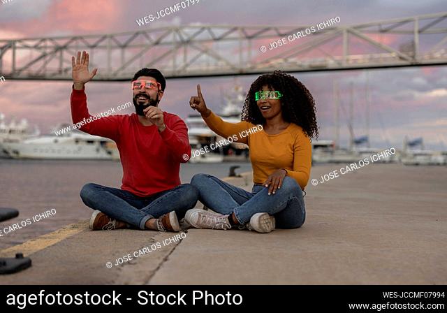 Happy couple gesturing with smart glasses sitting at promenade