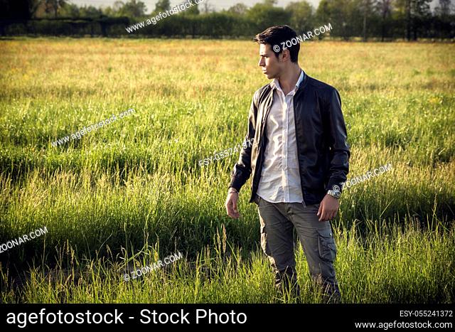 Handsome young man at countryside, in front of field or grassland, wearing white shirt and jacket, looking away in the distance