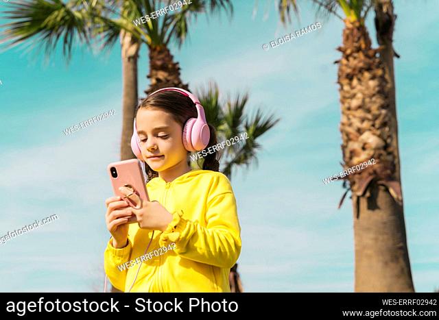 Portrait of little girl wearing yellow jacket listening music with headphones looking at smartphone