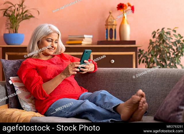 Old woman using mobile phone while relaxing on sofa in living room