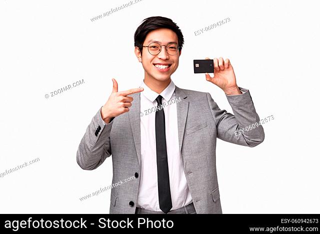 Waist-up portrait of satisfied, good-looking young assured asian man in suit, showing credit card, pointing at it with pleased smile
