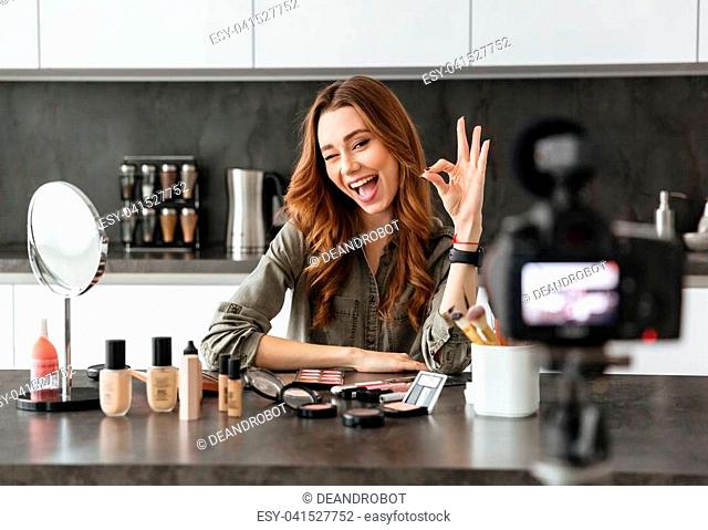 Smiling young girl recording her video blog episode about new cosmetic products while sitting at the kitchen table at home and applying make-up and showing ok...