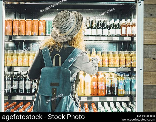 Back view of woman tourist with backpack in front of a window store full of drinks bottles choosing one to buy and drink