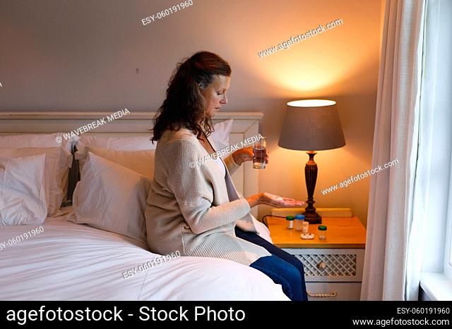 Senior caucasian woman in bedroom sitting on bed, holding pills