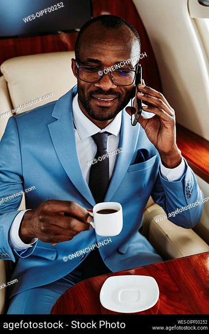 Young businessman with coffee cup talking on mobile phone in airplane