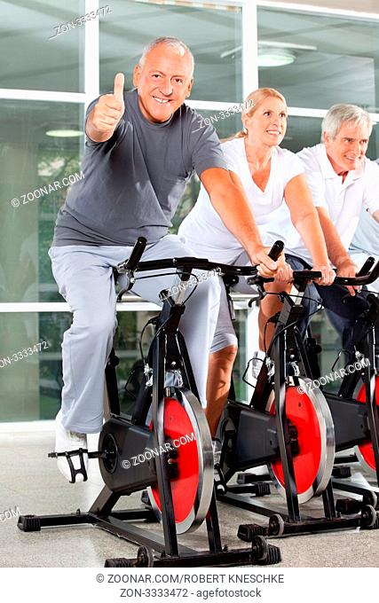 Lachender Senior auf Spinning-Rad im Fitnesscenter zeigt mit seinem Daumen nach oben