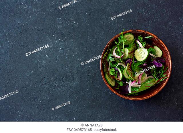 Fresh lettuce, arugula, frisee, basil, cucumber and onions salad in a clay bowl over black stone background. Vegan food. Top view. Copy space