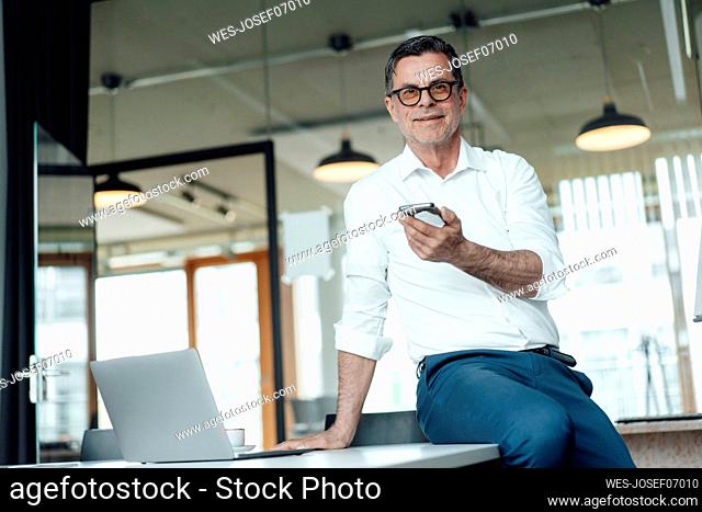 Smiling businessman with smart phone sitting on table at office