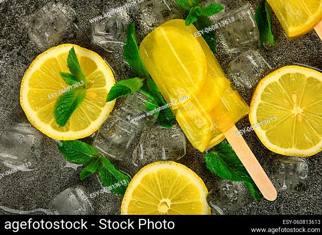 Close up frozen fruit juice popsicle with fresh lemon slices, green mint leaves and ice cubes on gray table surface, elevated top view, directly above