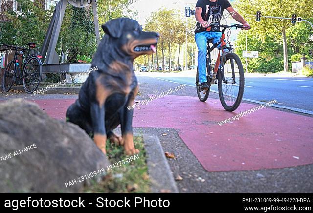 PRODUCTION - 06 October 2023, Baden-Württemberg, Esslingen am Neckar: A barking plastic dog stands on a sidewalk while a cyclist passes by on the sidewalk...