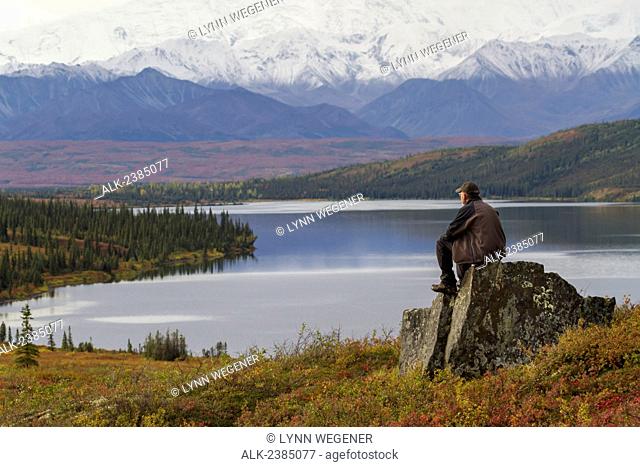 A male hiker rests on a large rock overlooking Wonder Lake in front of Mt. McKinley, Fall, Denali National Park, Interior Alaska, USA