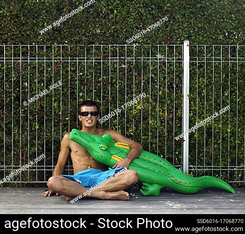 Young man sitting against pool fence holding crocodile blow up pool toy