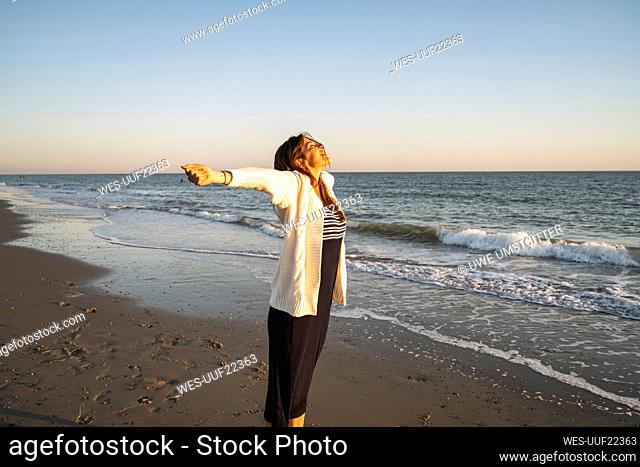 Carefree young woman standing at beach with arms outstretched during sunset