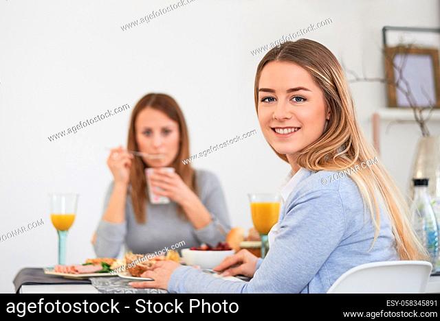 Pretty young teenage woman with a vivacious smile sitting at a table with her best friend enjoying breakfast and turning to smile at the camera