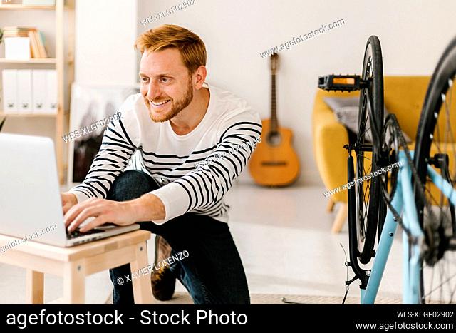 Smiling young man using laptop by bicycle in living room at home