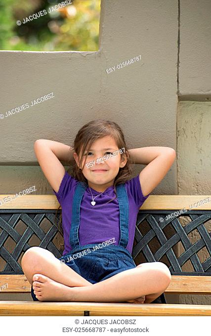 A young girl sits on an outside bench with heer legs crossed, her arms behind her head and her head against the wall, while giving a slight smile