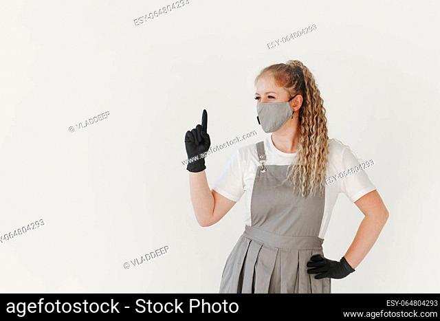 Woman beauty industry nail master in grey uniform and face mask and black gloves shows a sign with finger attention on white background