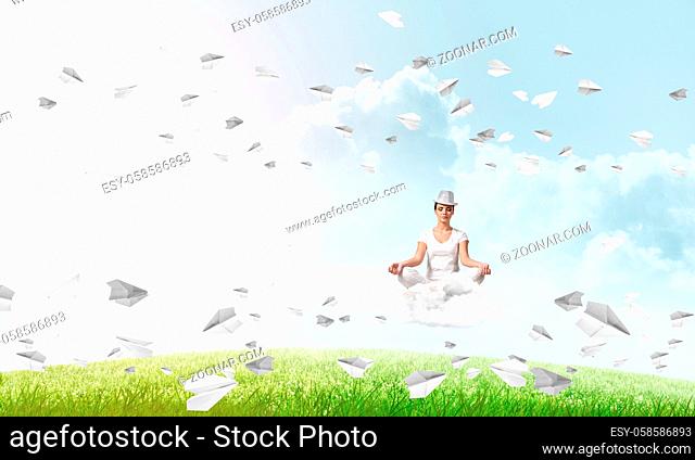 Young woman keeping eyes closed and looking concentrated while meditating on cloud among flying paper planes with bright and beautiful landscape on background
