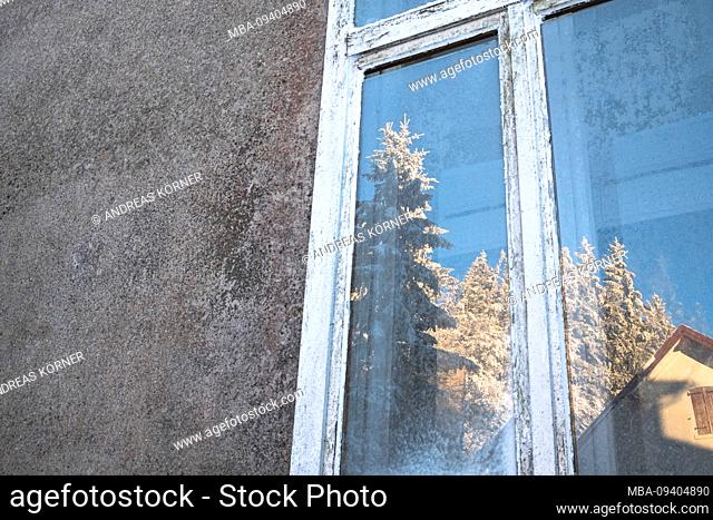 Snow-covered, old window with reflection of a sunny, wintry landscape and hut in gray facade in the Vosges, France