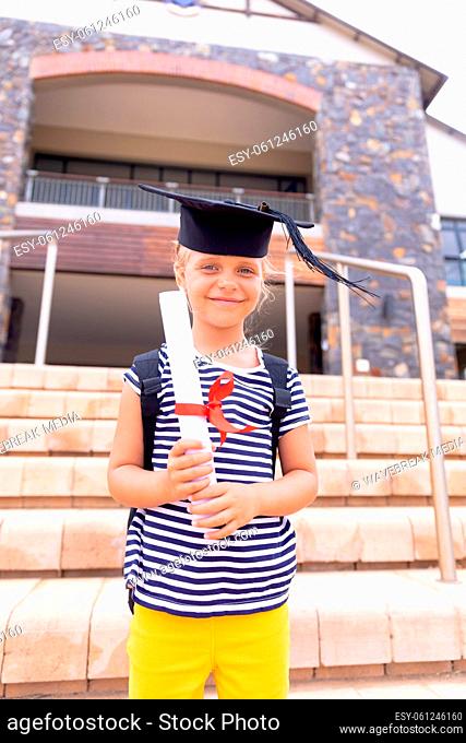 Portrait of smiling caucasian elementary schoolgirl with mortarboard degree standing against school