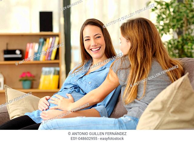 Happy pregnant woman talking with a friend who touches her belly sitting on a couch in the living room at home