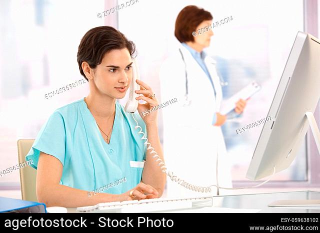 Medical assistant sitting at desk, talking on landline phone, using computer, doctor in background