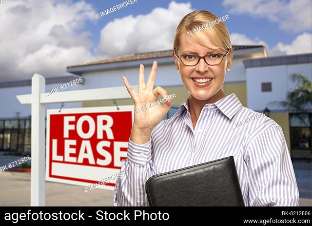 Smiling businesswoman with okay sign in front of vacant office building and for lease real estate sign