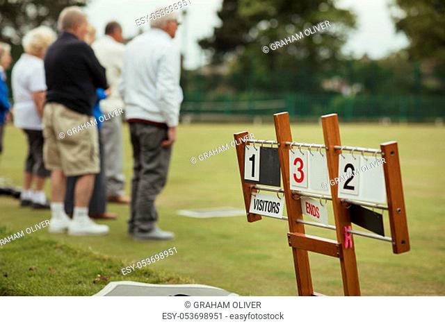 A close up shot of a lawn bowling scoreboard on grass, with senior adults spectating the lawn bowling game