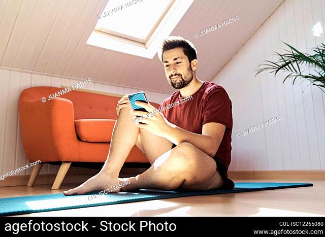 Man sitting on exercise mat, looking at phone