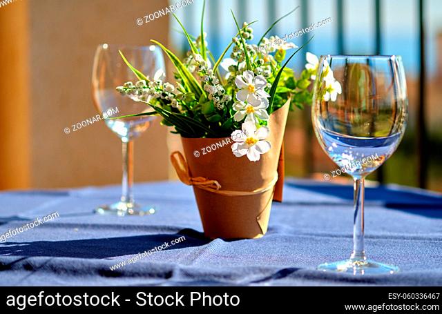 Two empty clean crystal wine glasses and artificial pretty blooming potted plant with flowers on table in summer terrace during sunny day warm weather close up...