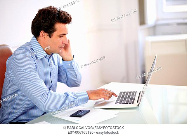 Portrait of a male hispanic boss working on his laptop and speaking on his mobile while sitting on manager desk