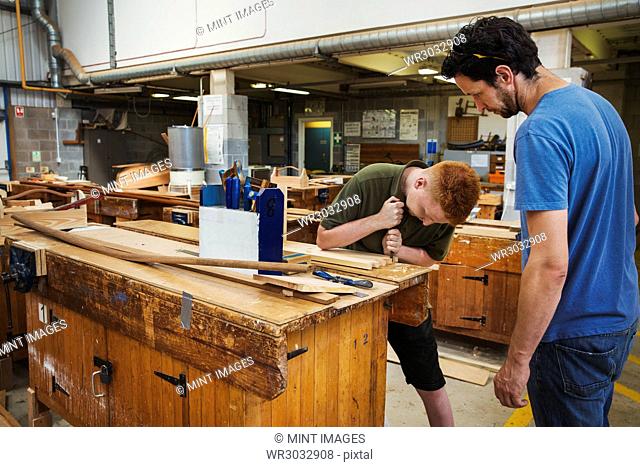 Two men standing at a workbench in a boat-builder's workshop, working on wooden joint