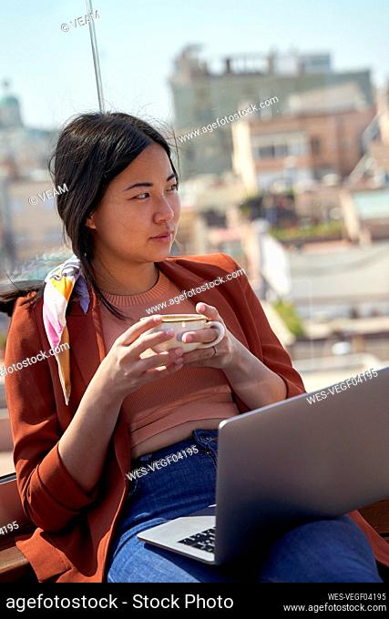 Thoughtful woman sitting with laptop and coffee cup at roof terrace