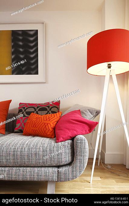 Large orange lampshade on tripod base standing at end of sofa covered in bright cushions. Lamp on | | Designer: Adrienne Chinn
