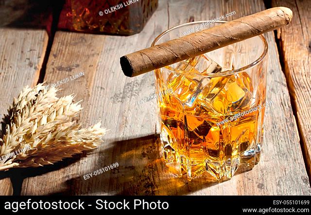 Glass of whiskey with ice and barley spikelets on wooden table