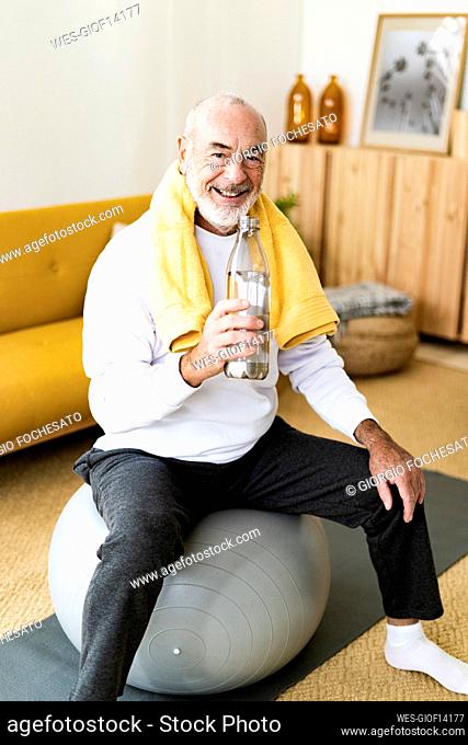 Smiling senior man holding water bottle on fitness ball at home