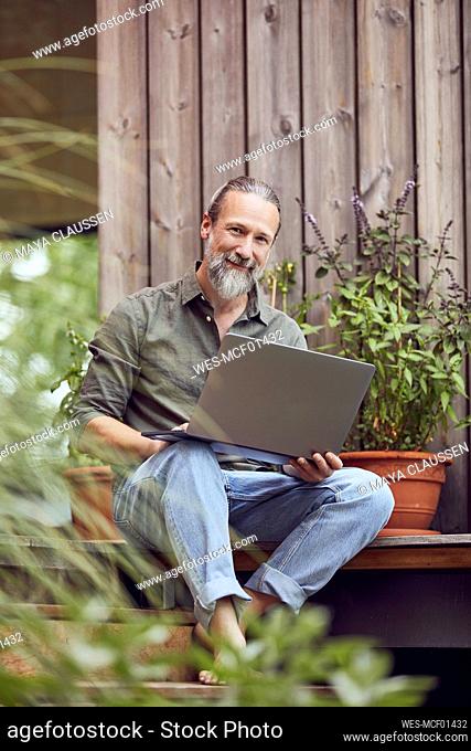 Smiling mature man with laptop sitting against tiny house
