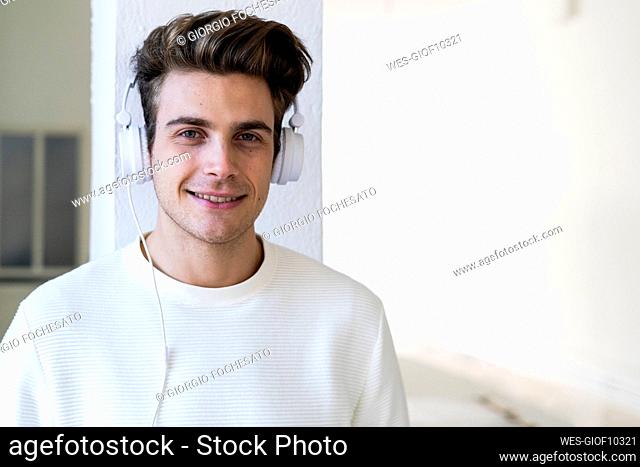 Smiling young man listening music through headphones against column in new home