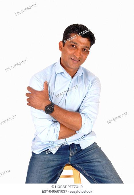 A handsome middle age East Indian man sitting on a bar chair in.jeans and a blue shirt, isolated for white background
