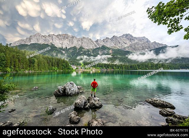 Young man standing on a rock on the shore, Eibsee lake in front of Zugspitze massif with Zugspitze, dramatic Mammaten clouds, Wetterstein range, near Grainau