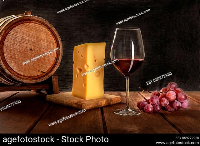 A glass of red wine with a wooden wine barrel, a large piece of Swiss cheese, and grapes on a dark rustic background, low key photo with copy space