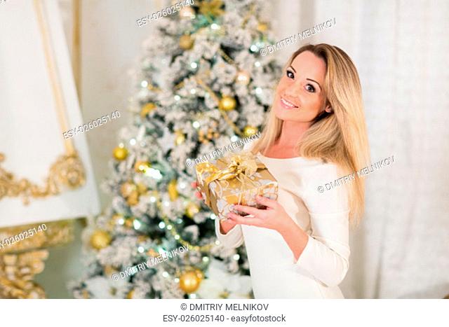 Young happy beautiful woman with gift box stands near Christmas tree in the room of the house. Merry Christmas and New Year
